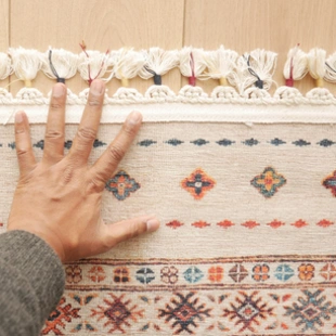 Illustration - Ihr Experte für schönes Wohnen - Eine Nahaufnahme einer Hand, die auf einem wunderschönen, farbenfrohen Vintage-Teppich aus der Vergangenheit platziert ist.