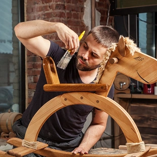 Schutzlasuren - Ein Holzschaukelpferd wird mit einem Pinsel mit Holzlasur Holzschutz transparent gestrichen.