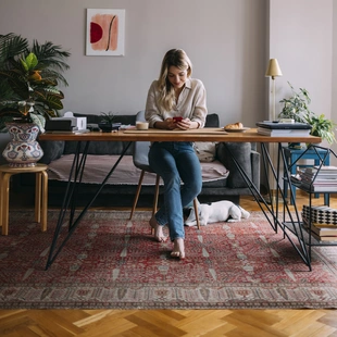 Illustration - Ihr Experte für schönes Wohnen - Schöne lächelnde Frau sitzt am Schreibtisch im Home Office unter dem Schreibtisch liegt ein Orientteppich.